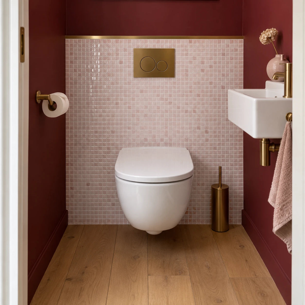 Toilettes avec WC suspendu, plaque de commande dorée et carrelage adhésif mosaïque rose poudré sous un mur bordeaux